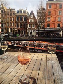 Im Hintergrund sind viele schmale bunte Häuser zu sehen und ein Boot in der Gracht. Im Vordergrund sehen 3 Weingläser auf einem hölzernen Tisch.